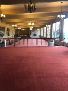 Clean red colored carpet in a bi-level large empty dining area with big windows on the right and a stone fireplace in the back with stacked tables and 2 garbage cans.