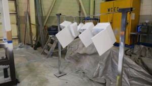 5 metal cabinet components primed in white paint hung to dry on a metal line structure within a manufacturing environment.